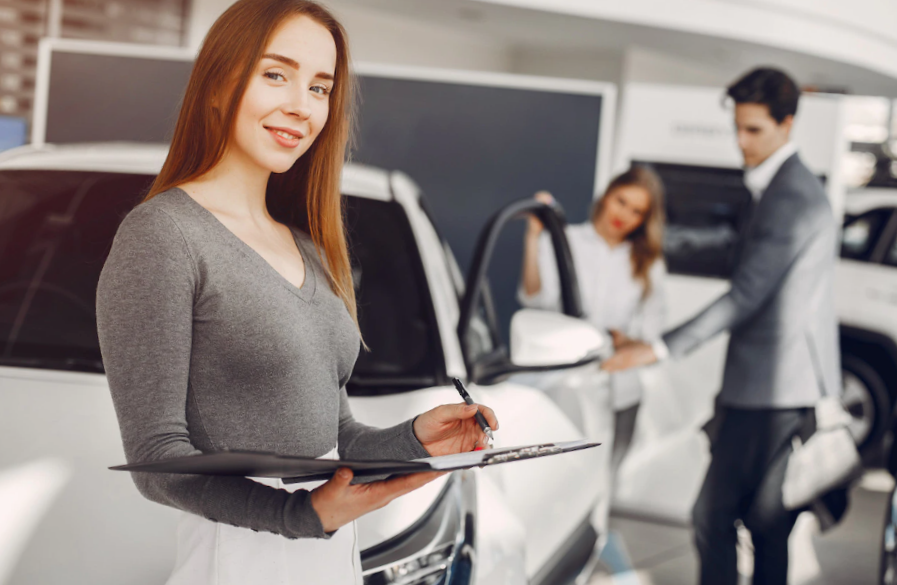 smiling woman with clipboard and pen in hands and male coworker showing a car to a female customer behind